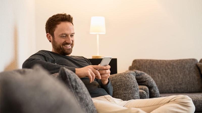 Mand slapper af på grå sofa med smartphone, hvid gulvlampe i baggrunden, hyggelig stue