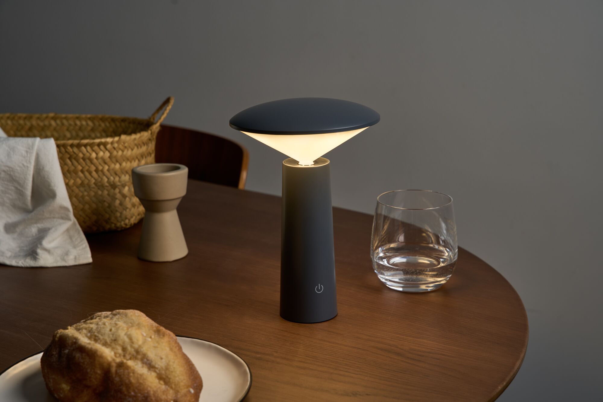 Modern black metal table lamp with warm white light on wooden table near glass and bread basket