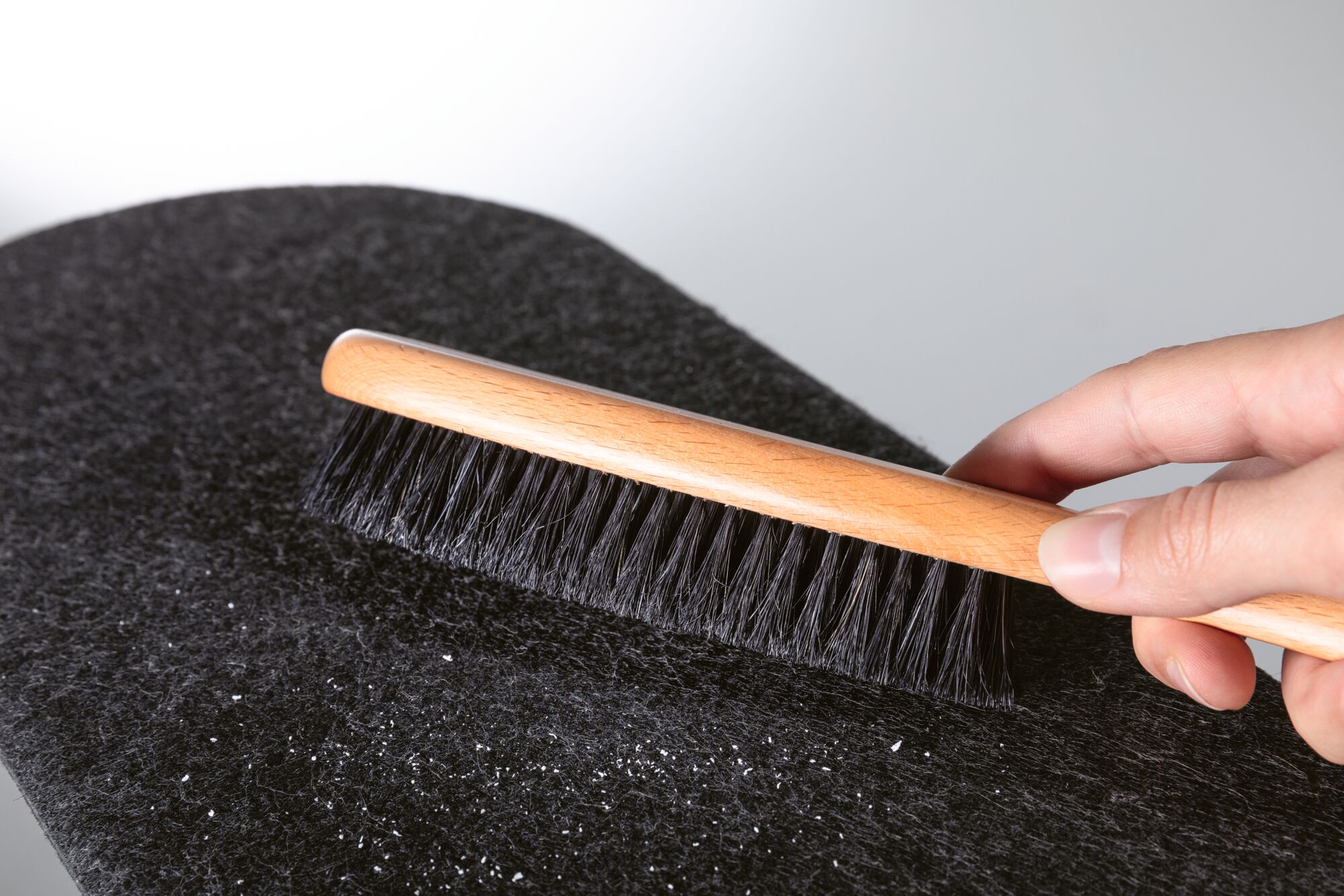 Hand holding wooden brush with black bristles cleaning black felt fabric from dust and lint.