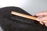 Hand holding wooden brush with black bristles cleaning black felt fabric from dust and lint.