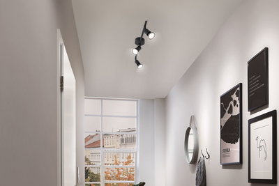 Black LED ceiling light with three adjustable spotlights in modern hallway with wall decor and window.