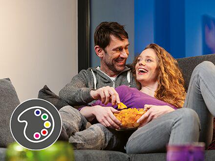 Couple relaxing on sofa with chips, icon for eye-friendly LED color rendering and lighting.