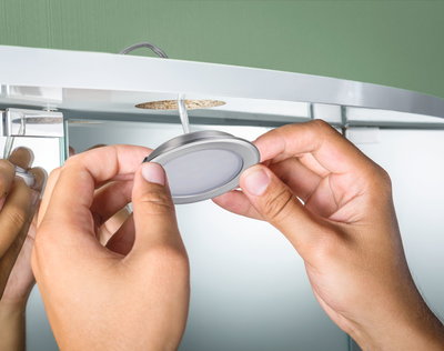 Hands installing round brushed aluminum LED recessed light in furniture with green wall background