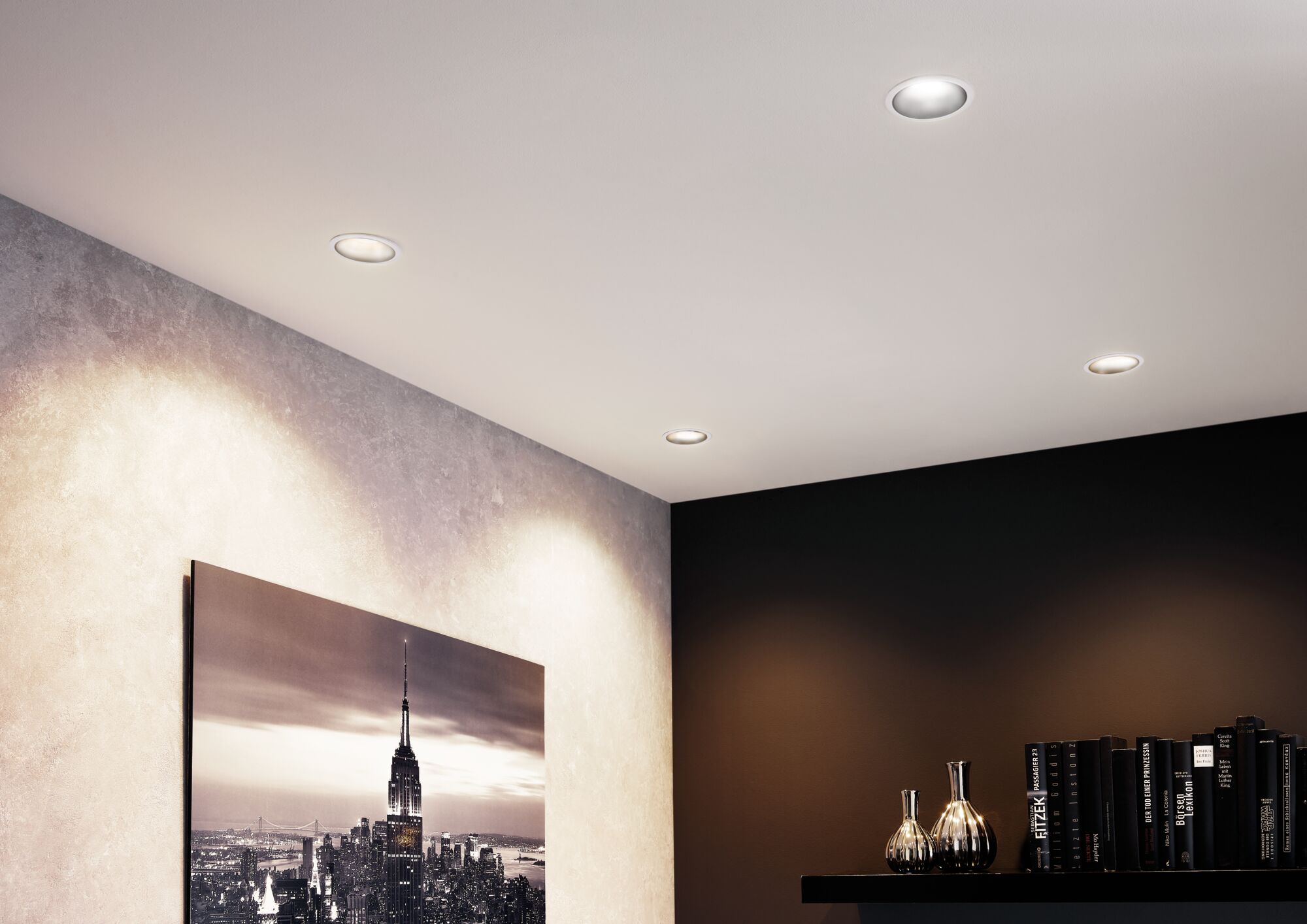 Vier weiße LED-Einbaustrahler in Decke beleuchten modernes Wohnzimmer mit Kunst und Bücherregal.