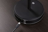 Black round lamp base with white stitching and USB-C charging cable on dark wood surface