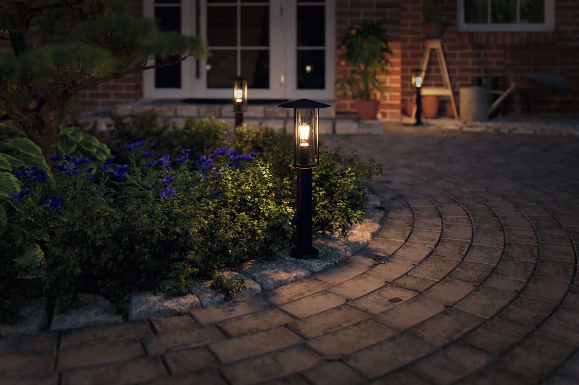 Zwarte metalen buitenlamp met warm wit licht langs tuinpad bij bloeiende planten.