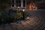 Schwarze Außenleuchte aus Metall mit warmweißem Licht im Garten neben lila Blumen und Steinweg