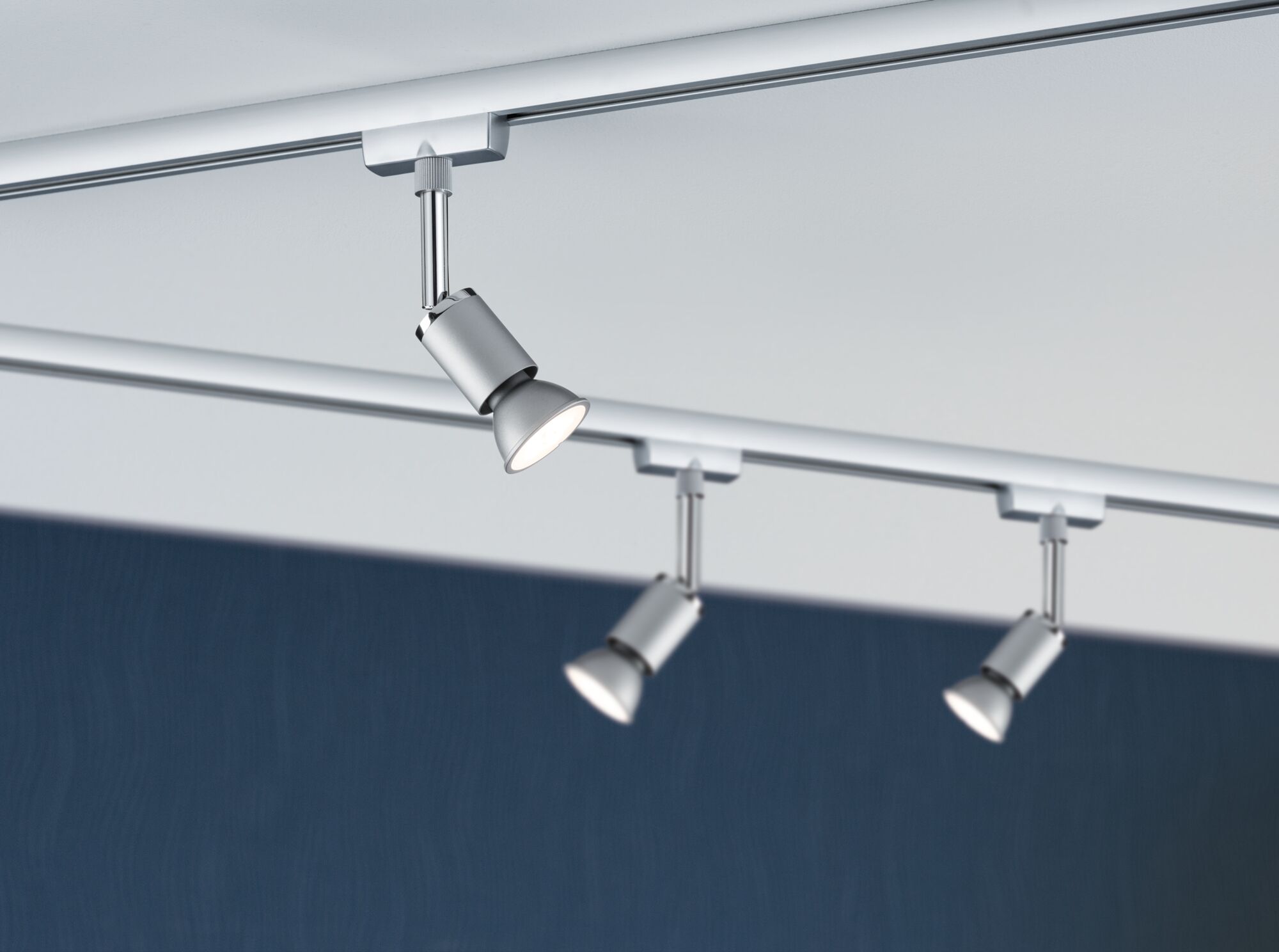 Tre sølvfarvede LED-spotlights i metal på skinnesystem mod hvidt loft og blå baggrund