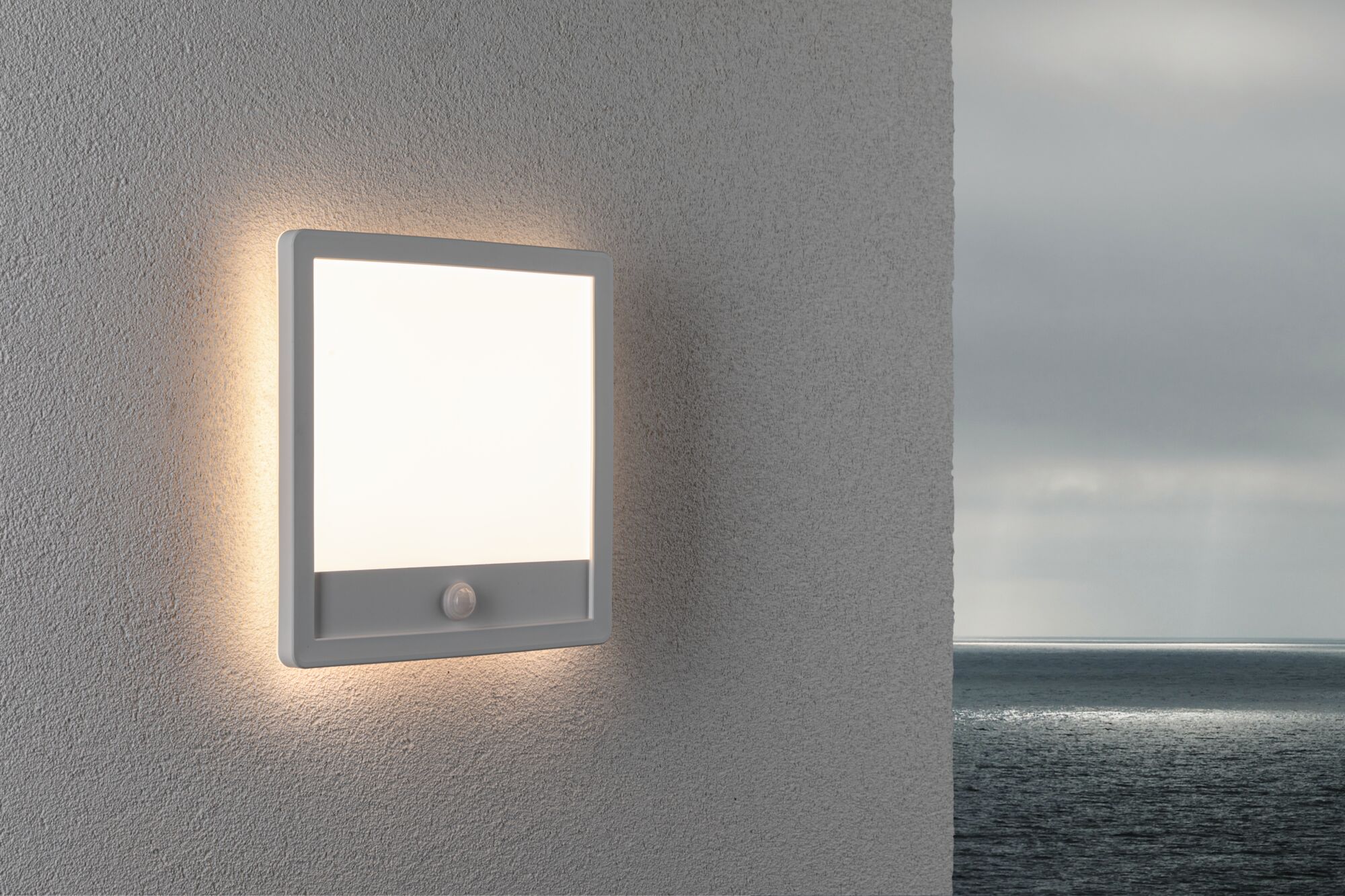 Square white LED wall light with motion sensor on grey exterior wall overlooking the sea