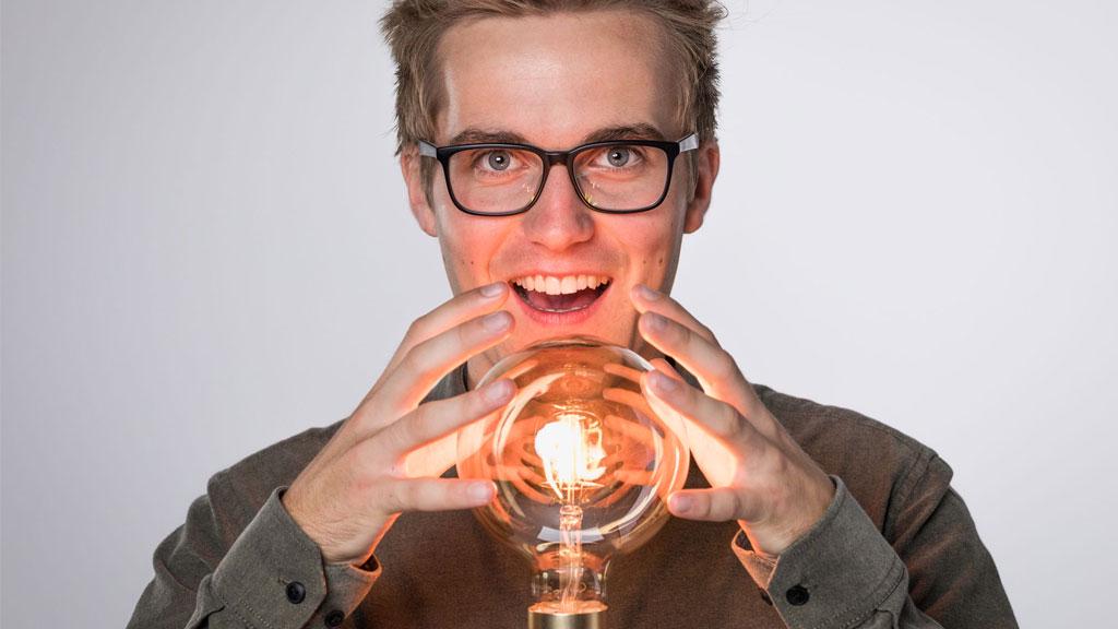 Junger Mann mit Brille hält eine leuchtende Glühbirne aus Glas, Symbol für Innovation und Lichttechnik.
