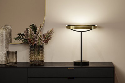 Moderne Tischleuchte mit goldfarbenem Metallring und schwarzem Ständer auf schwarzem Sideboard mit Vase.
