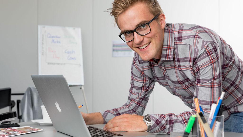 Junger Mann mit Brille arbeitet an silbernem Laptop in modernem Büro mit Flipchart im Hintergrund