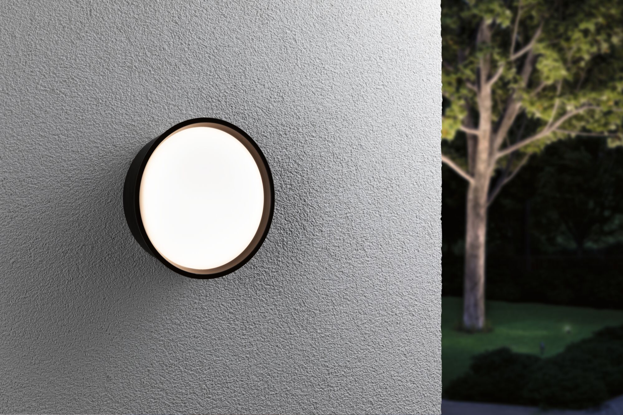 Round outdoor wall light with black frame and warm white glow on grey wall in garden