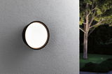 Round outdoor wall light with black frame and warm white glow on grey wall in garden