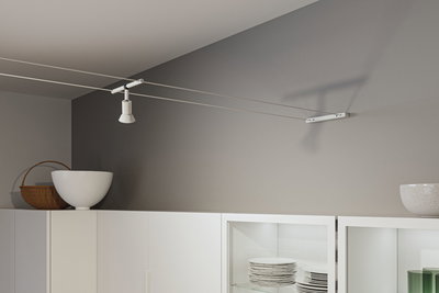 Modern white metal cable spotlight on grey wall above white kitchen cabinet with dishes.