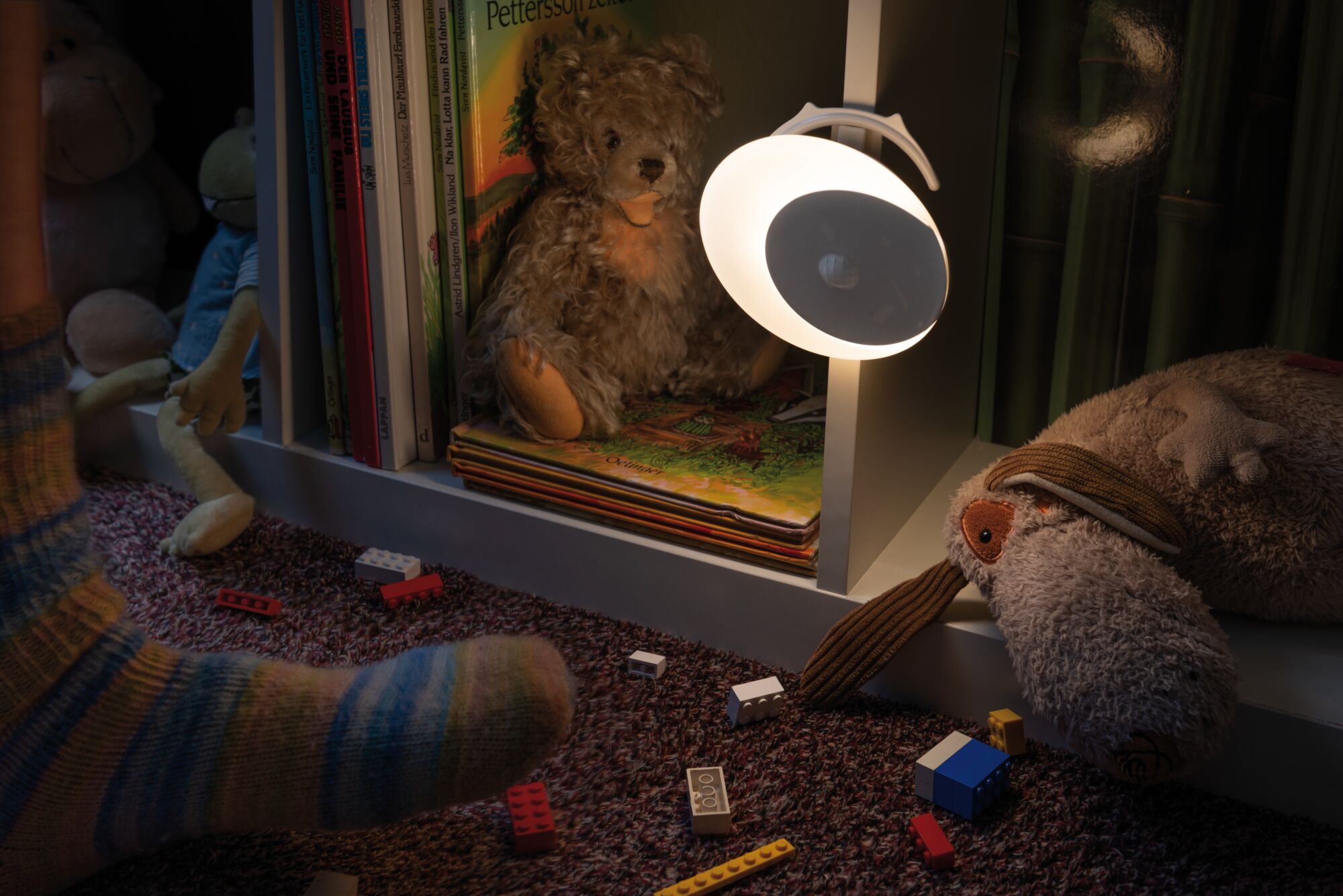 Weiße LED-Nachtlampe mit Bewegungsmelder an weißem Regal neben Kuscheltieren und Kinderbüchern.