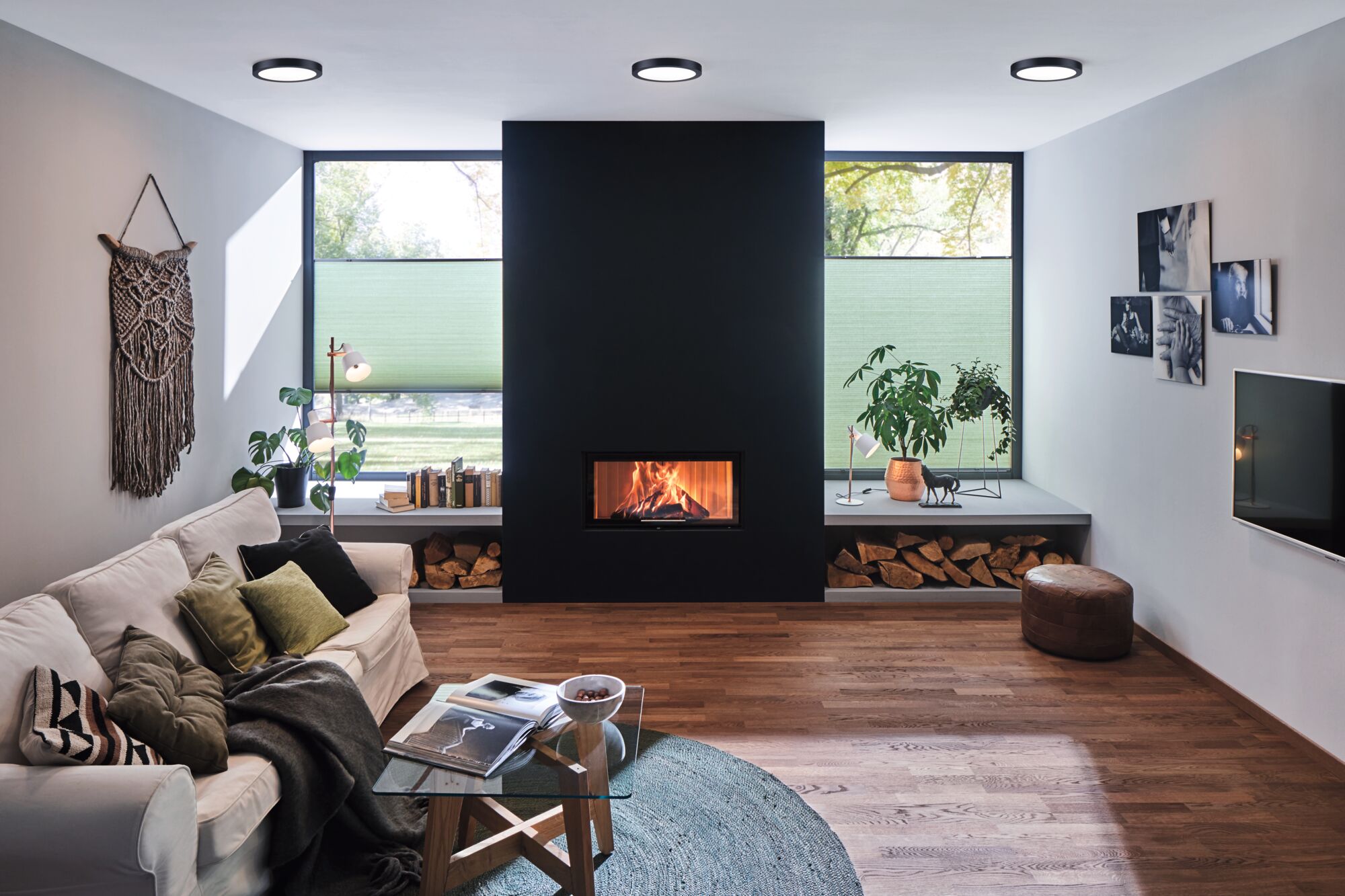 Modernes Wohnzimmer mit schwarzem Kamin, Holzlager, weißen Deckenleuchten und gemütlicher Einrichtung mit Sofa und Pflanzen.