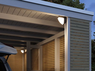 Modern outdoor wall light with white illumination on wood paneling and carport in gray and natural wood.
