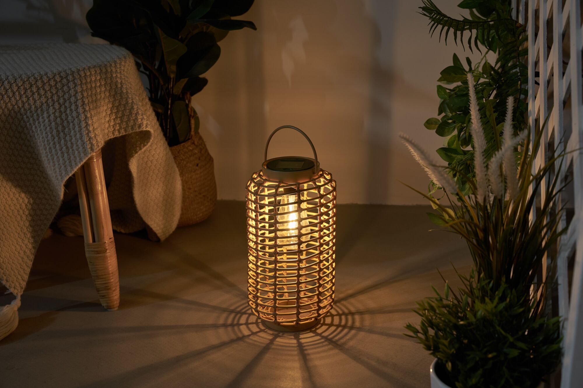 Warm light from a round lantern made of woven natural material on floor with plants in background.