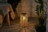 Warm light from a round lantern made of woven natural material on floor with plants in background.