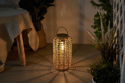 Warm light from a round lantern made of woven natural material on floor with plants in background.