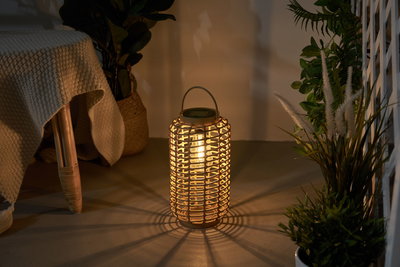 Warm light from a round lantern made of woven natural material on floor with plants in background.