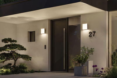 Modern white outdoor wall light with warm white glow at entrance featuring black door and greenery.