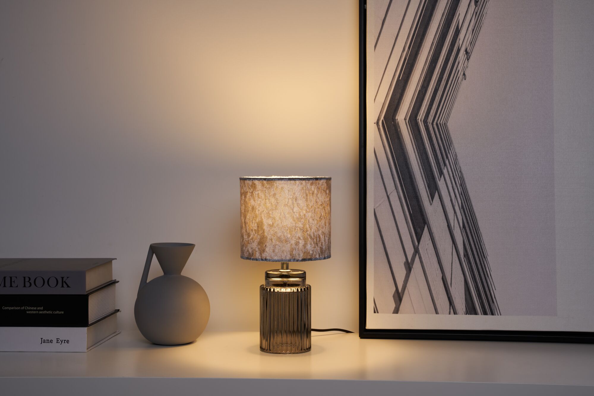 Moderne Tischlampe mit grauem Stoffschirm und geriffeltem Glasfuß auf weißem Regal mit Deko.