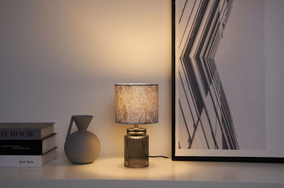 Moderne Tischlampe mit grauem Stoffschirm und geriffeltem Glasfuß auf weißem Regal mit Deko.
