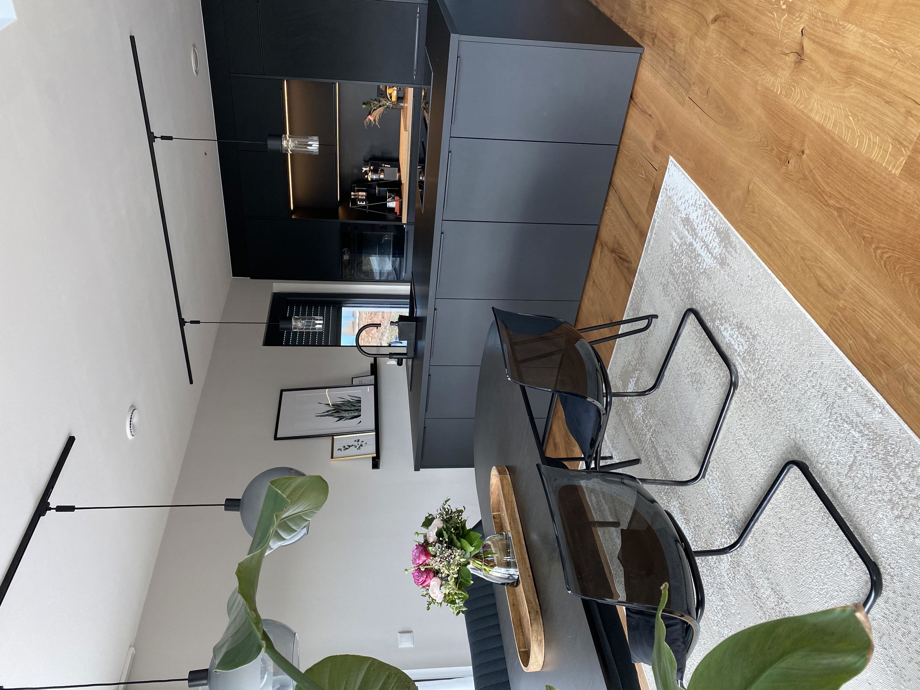 Moderne Küche mit mattschwarzen Schränken, Holzfußboden und transparenten Stühlen auf hellem Teppich.