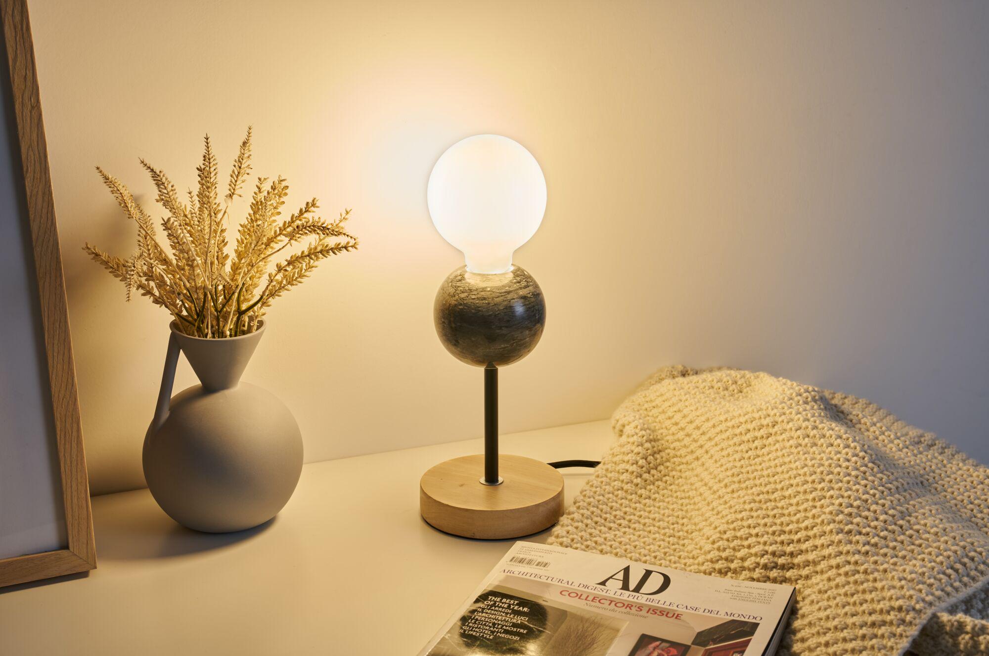 Modern table lamp with wooden base and black metal stem, frosted glass globe emits warm light indoors.