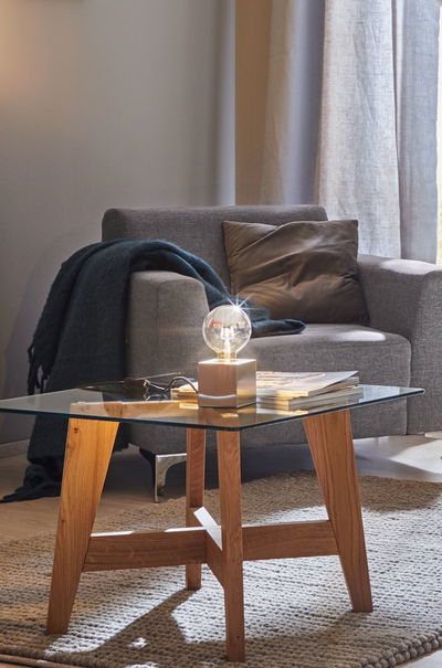 Table d'appoint moderne avec plateau en verre et cadre en bois, décorée d'une ampoule et de magazines dans le salon.