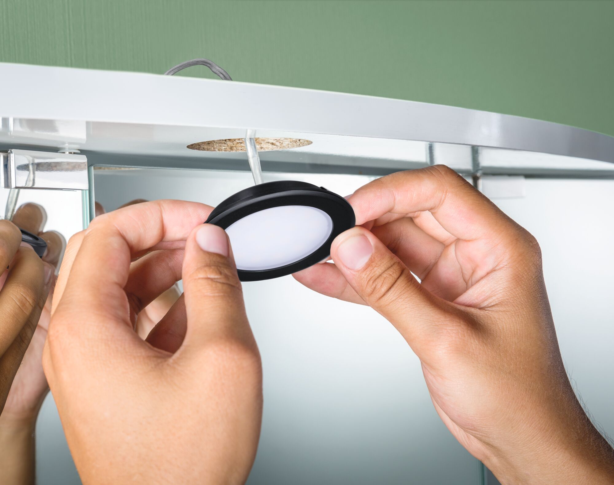 Main installant une lampe LED ronde noire sous meuble avec diffuseur blanc pour éclairage mobilier