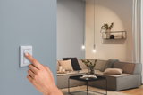 Hand bedient witte wandschakelaar op blauwe muur in moderne woonkamer met grijze bank en hanglampen