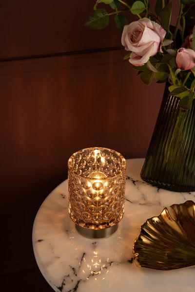 Glass tealight holder with round embossing and warm light on marble table near vase with pink roses.