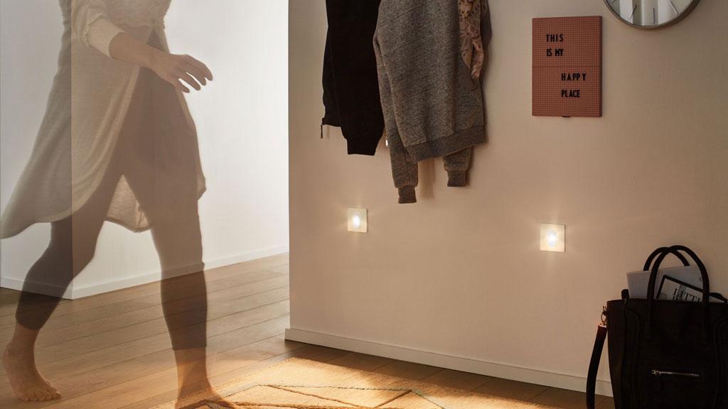 Hallway with two white LED motion sensors mounted on wall, warm lighting and coats hanging on hooks.