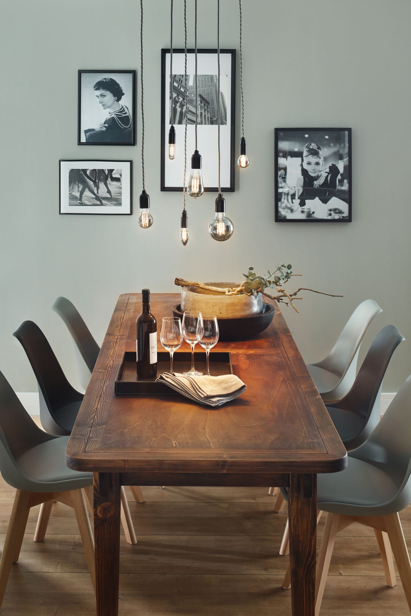Table à manger en bois avec six chaises grises et ampoules LED filament en verre fumé suspendues.