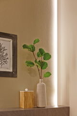 Beige ceramic vase with green leaves beside wooden box on grey shelf against lit wall