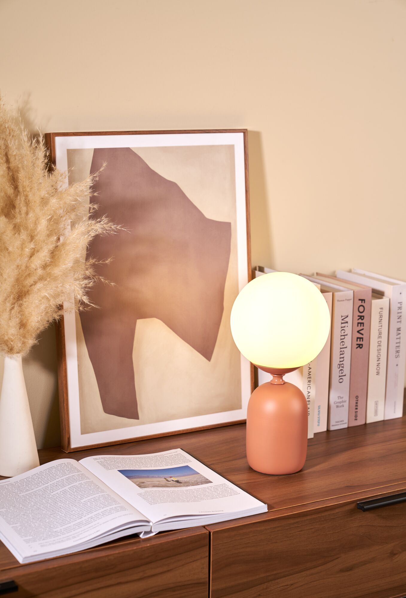 Modern table lamp with round frosted glass and matte terracotta ceramic base on wooden sideboard with books.