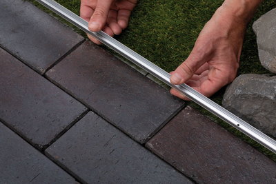 Profilé en aluminium posé entre pavés foncés et gazon synthétique vert comme bordure.