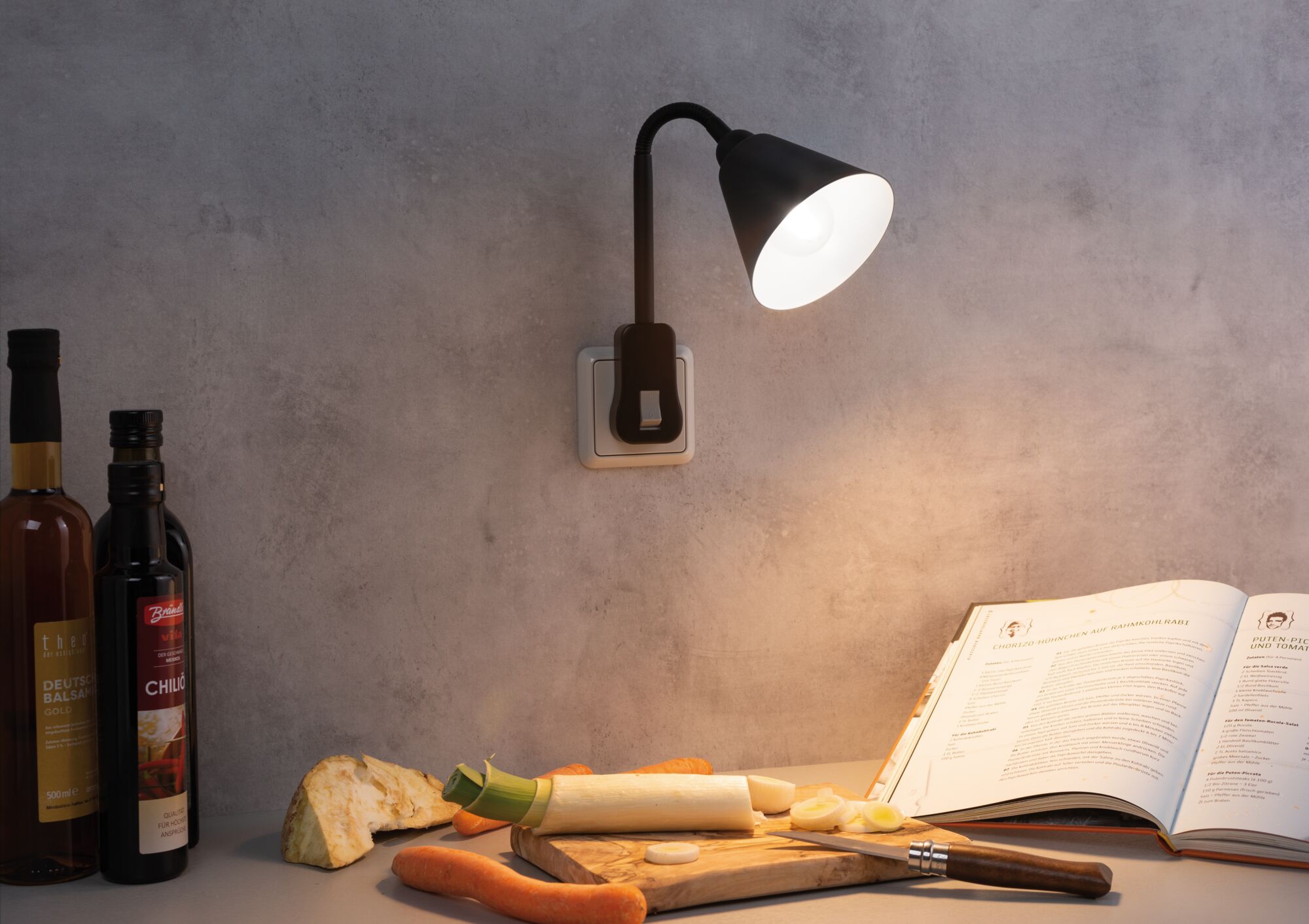 Black wall lamp with flexible arm on grey wall lighting cookbook and vegetables on wooden board.