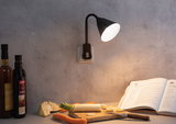 Applique murale noire à bras flexible sur mur gris éclairant un livre de cuisine et des légumes sur planche en bois.