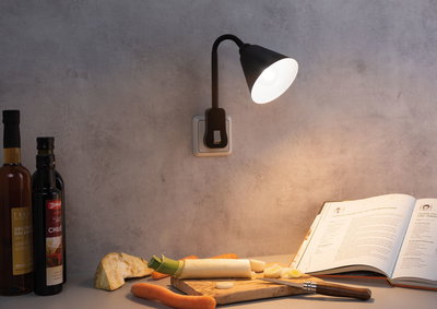 Zwarte wandlamp met flexibele arm aan grijze muur verlicht kookboek en groenten op houten plank.