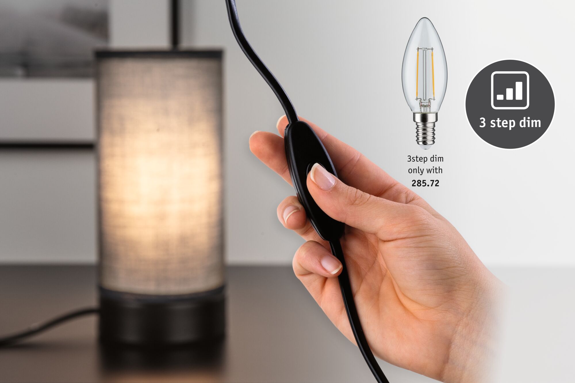 Hand operating black dimmer switch in front of lit table lamp with fabric shade for ambient lighting control.