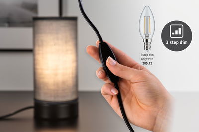 Hand operating black dimmer switch in front of lit table lamp with fabric shade for ambient lighting control.