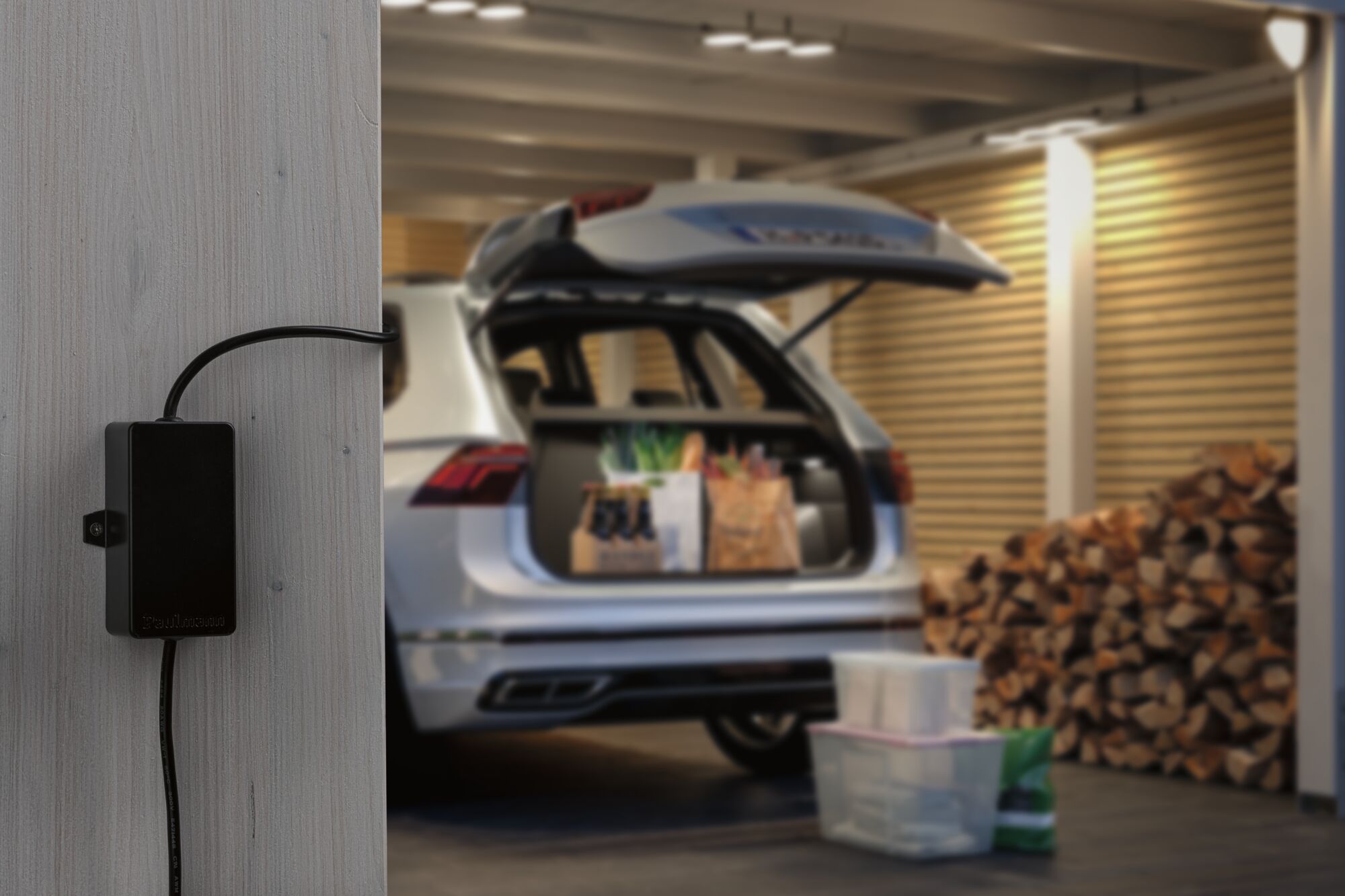 Black Paulmann cable connection box on grey wooden wall in garage with open car trunk and stacked firewood.
