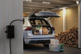 Zwart Paulmann kabelaansluitdoosje aan grijze houten wand in garage met open kofferbak en gestapeld brandhout.