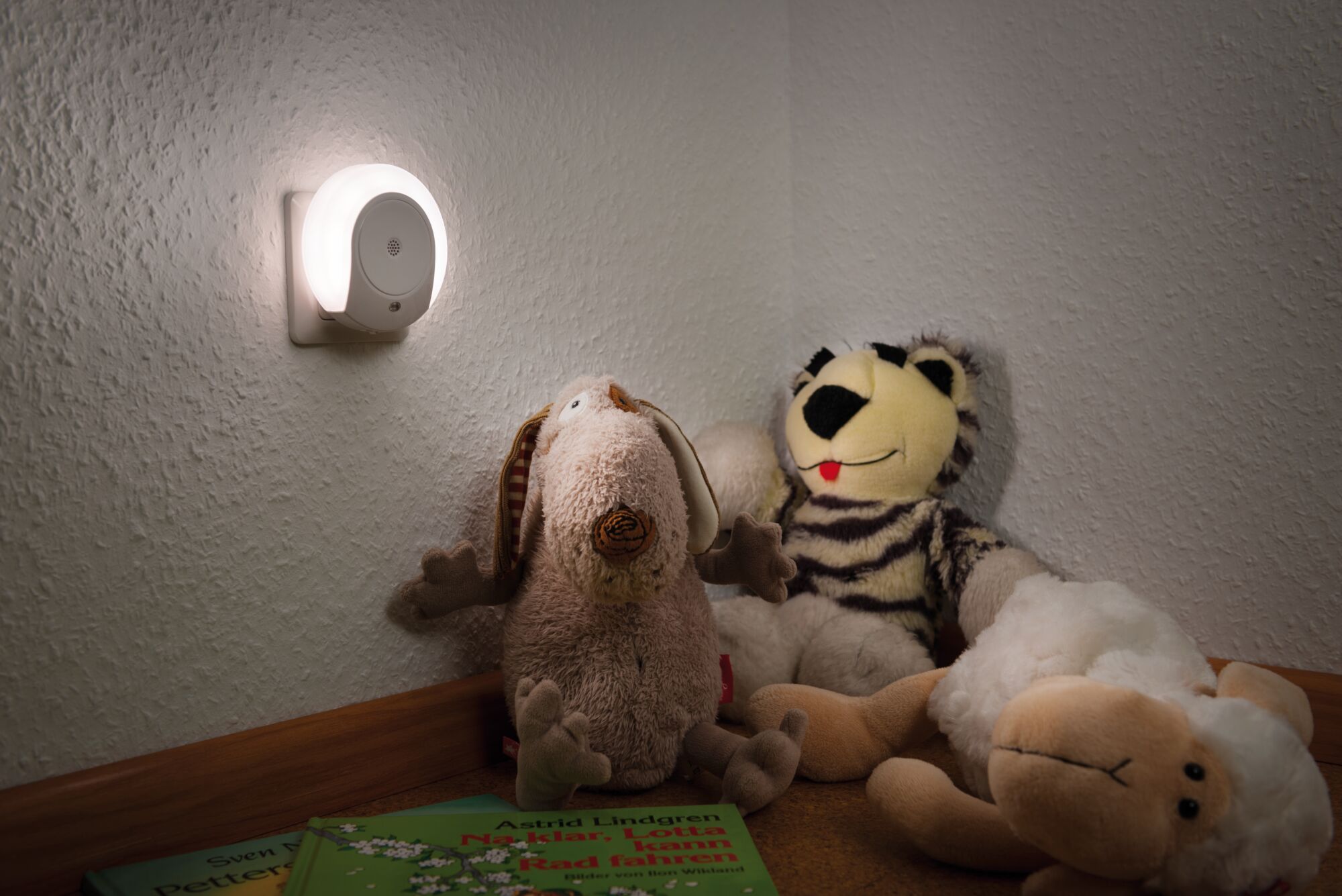 White LED night light plug on wall illuminating stuffed animals and children's books in room.