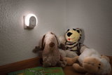 White LED night light plug on wall illuminating stuffed animals and children's books in room.