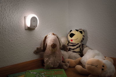 White LED night light plug on wall illuminating stuffed animals and children's books in room.